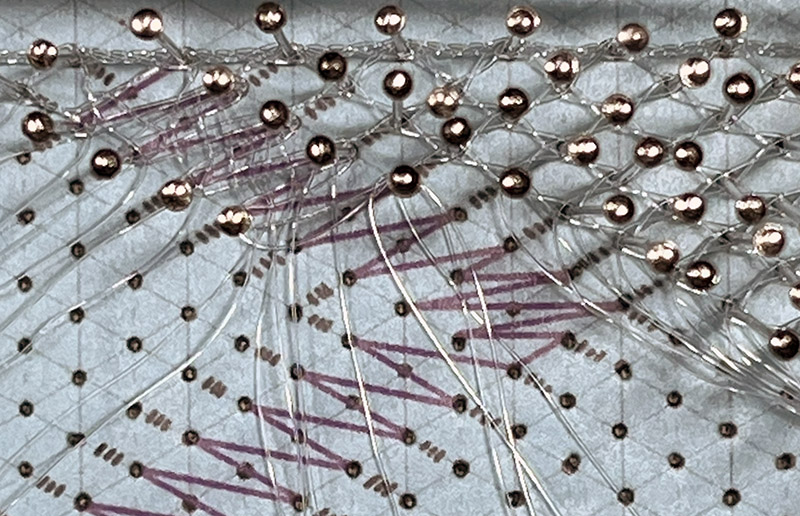 Close-up of bobbin lace in progress using clear fishing line, lit with regular white light.